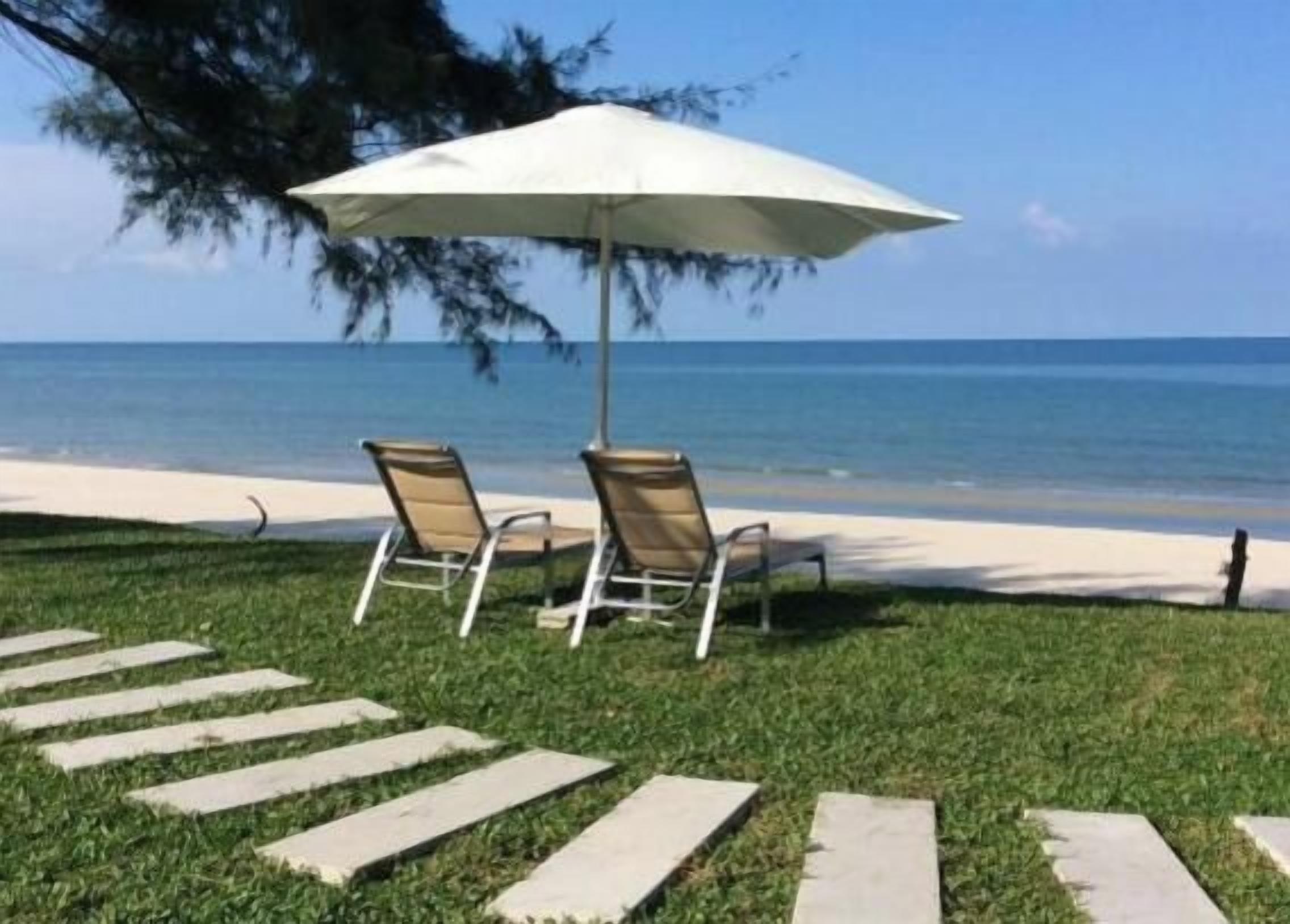 Deux fauteuils de jardin sont placés sous un parasol blanc, sur une pelouse. Un chemin de pierres mène à la plage.