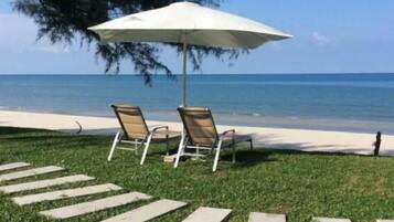 Deux fauteuils de jardin sont placés sous un parasol blanc, sur une pelouse. Un chemin de pierres mène à la plage.