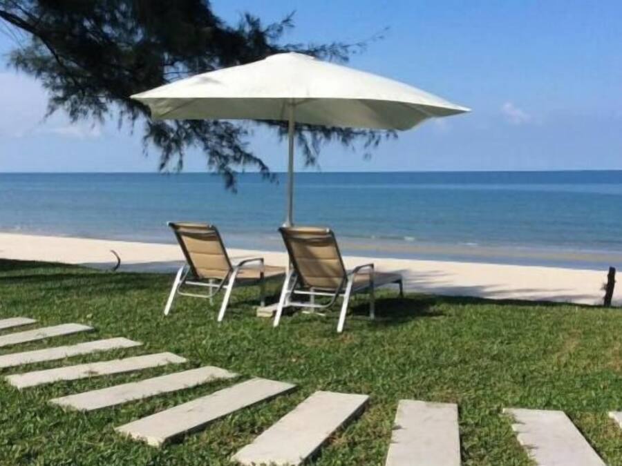 Twee lounge stoelen onder een witte parasol op een gazon, met een pad van stenen dat naar het strand leidt.