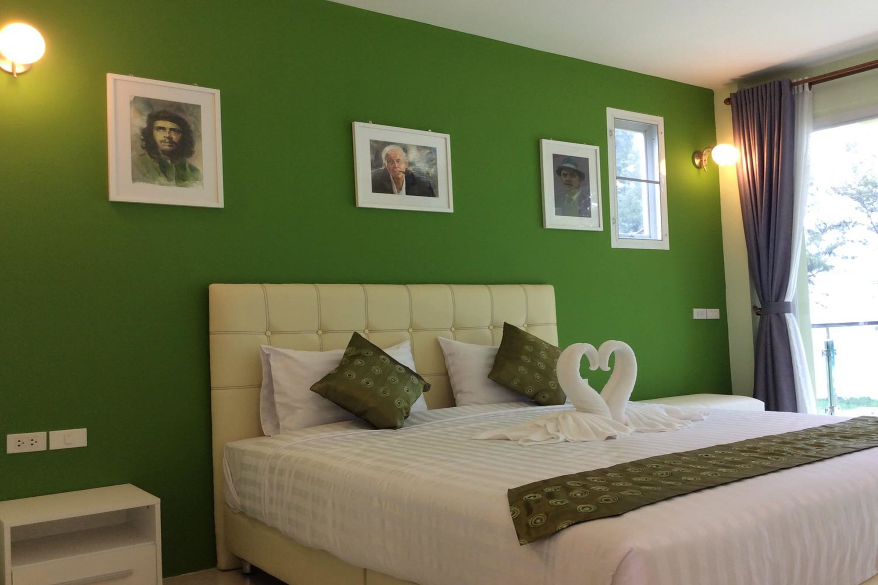 A neatly made bed with white linens and decorative pillows, a green wall with framed pictures, and a window with curtains.