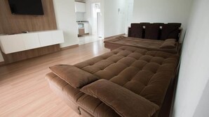 A modern living room with a large brown sofa, a flat-screen TV mounted on the wall, and a kitchen area in the background. - Residencial Scheifler (Gramado)
