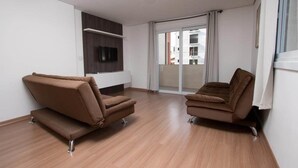 A modern living room with two brown sofas, wooden flooring, a flat-screen TV mounted on the wall, and a large sliding glass door leading to a balcony. - Residencial Scheifler (Gramado)