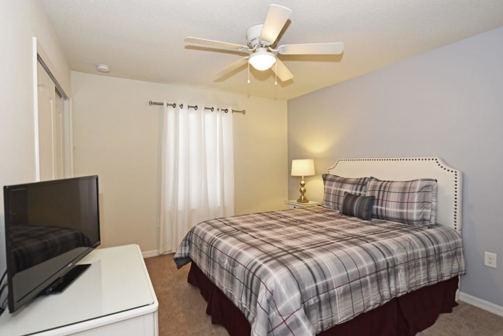 A bedroom with a bed, a TV on a stand, a ceiling fan, and a nightstand with a lamp.