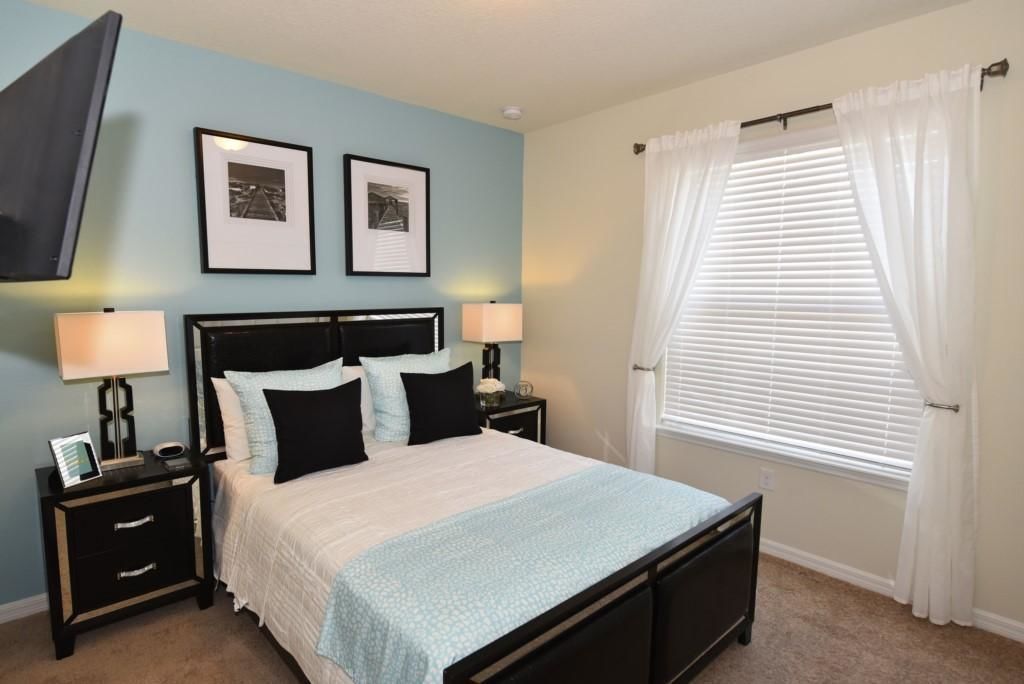 A bedroom with a bed, two bedside tables, a nightstand, a TV mounted on the wall, and a window with white curtains.