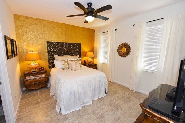 A bedroom with a bed, two bedside tables with lamps, a ceiling fan, a wall-mounted decorative piece, and a television.
