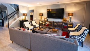 A modern living room with a sectional sofa, two armchairs, a coffee table, and a TV. A staircase is visible in the background. - 216 Watersong House 7 Bedroom by Florida Star (Davenport)