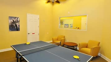 Una habitación con mesa de ping pong, dos sillones amarillos, una mesita redonda y un ventilador de techo.