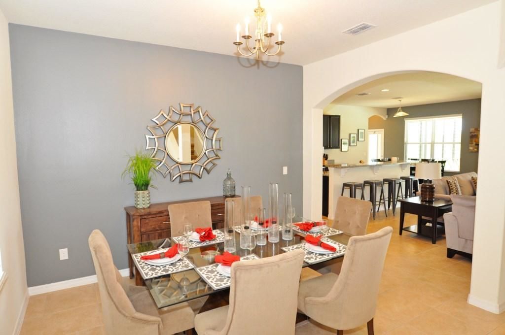 A dining area with a glass table set for six, surrounded by beige chairs. A decorative mirror hangs on the wall, and a chandelier is visible above.