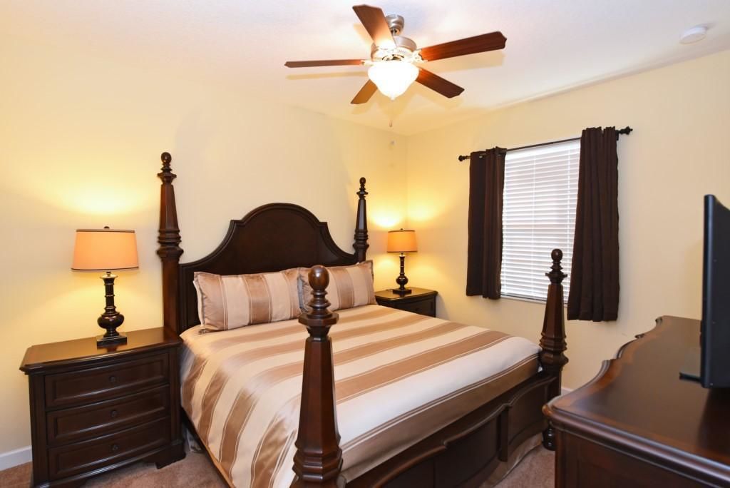 A bedroom with a four-poster bed, two bedside tables with lamps, a ceiling fan, and a window with curtains.