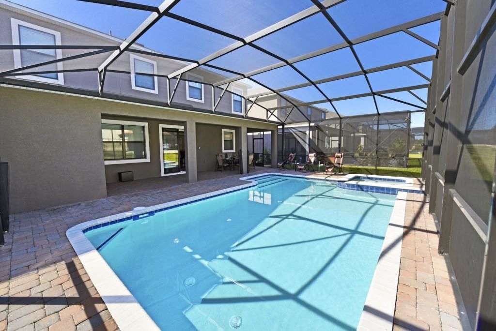 A swimming pool with a glass ceiling structure, surrounded by a brick patio and a building with large windows.