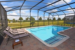 A covered swimming pool with lounge chairs and a table, surrounded by a metal fence and brick flooring.