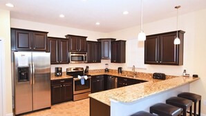 A modern kitchen with dark cabinetry, stainless steel appliances, granite countertops, and bar stools. - 2383 Providence House 6 Bedroom by Florida Star (Davenport)
