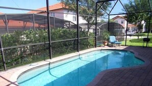 Une piscine avec une terrasse en briques, entourée d’une structure en verre, et un ensemble de chaises et de table installé à proximité.