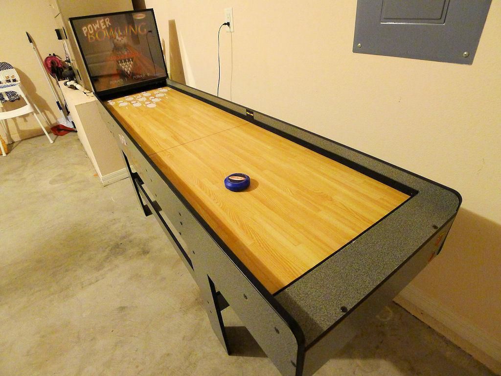 Een shuffleboardtafel met een blauwe schijf in het midden, een televisiescherm waarop 'Power Bowling' te zien is op de achtergrond, en een witte stoel aan de linkerkant.