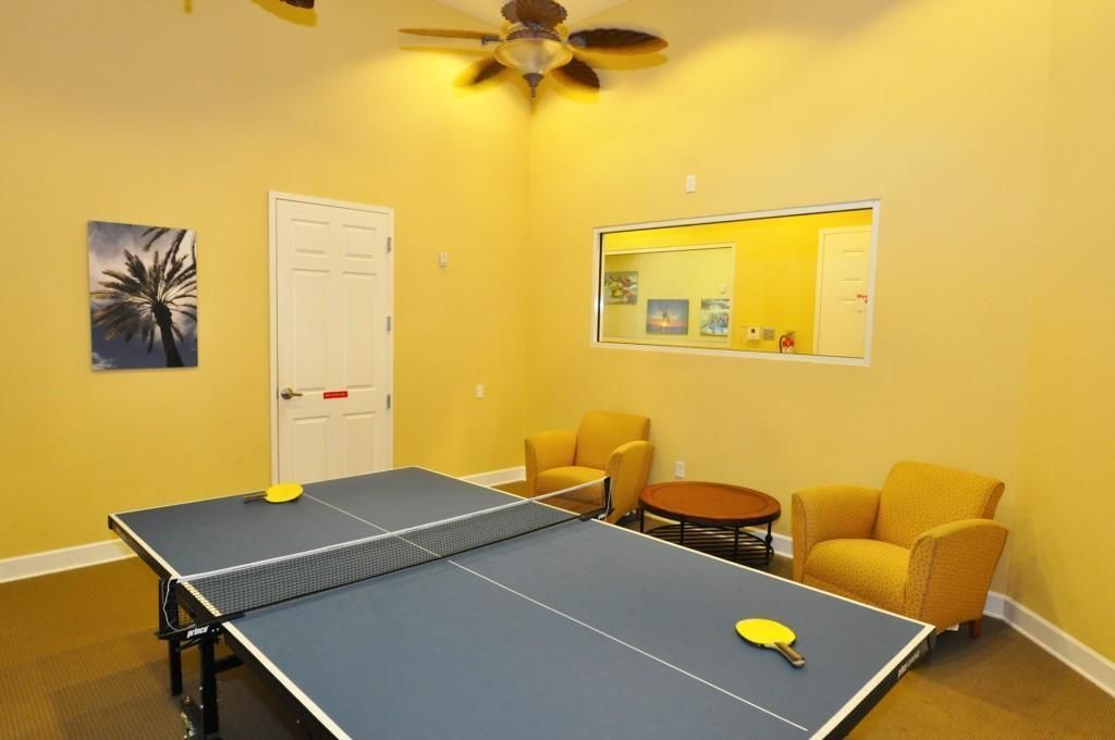 Ein Zimmer mit einem Tischtennisplatte, zwei gelben Sesseln, einem kleinen runden Tisch und einem Deckenventilator.