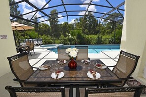 Et udendørs spiseområde med udsigt til poolen, med et bord dækket til fire personer, tallerkener, glas og en blomstervase.