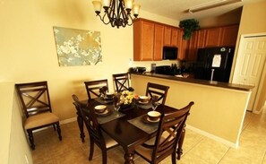 A dining area with a dark wooden table and chairs, a chandelier, and a kitchen in the background. - 2755 Oakwater Condo 2 Bedroom by Florida Star (Kissimmee)