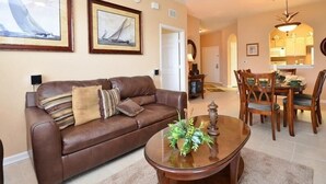 A living room with a brown leather sofa, a glass coffee table with a floral centerpiece, and a dining area with wooden chairs and a table. - 2809 Windsor Hills Condo 3 Bedroom by Florida Star (Kissimmee)