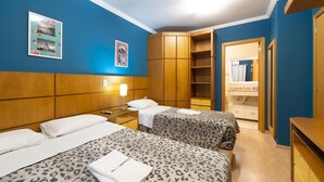Quarto de hotel com duas camas, um guarda-roupa, um espelho e uma pequena escrivaninha com abajur.