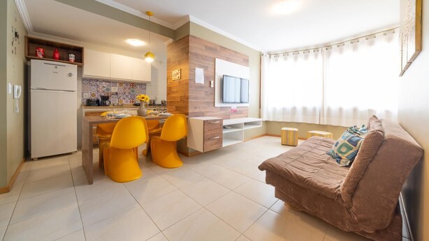 Uma sala de estar moderna com um sofá marrom, cadeiras amarelas, uma cozinha com uma geladeira branca e uma TV fixada na parede.