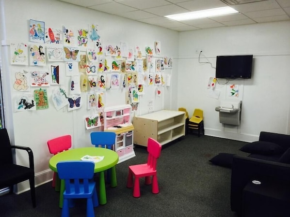 Una sala de juegos infantiles con sillas coloridas, una mesa y paredes decoradas con dibujos de niños.