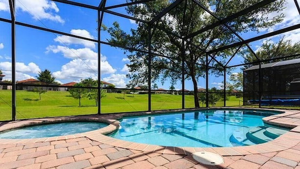 A swimming pool with a brick deck, surrounded by a glass enclosure with a view of a green lawn and houses. - 320 High Grove House 5 Bedroom by Florida Star (Clermont)