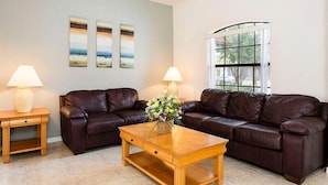A living room with two brown leather sofas, a wooden coffee table, and two lamps. There are three framed pictures on the wall and a window with a view of greenery. - 320 High Grove House 5 Bedroom by Florida Star (Clermont)