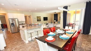 A modern kitchen with a central island, dining area with red chairs, and a large window with curtains. - 394 Watersong House 6 Bedroom by Florida Star (Davenport)