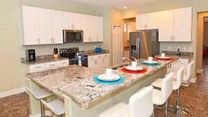 A modern kitchen with white cabinets, granite countertops, a central island with bar stools, and stainless steel appliances. - 394 Watersong House 6 Bedroom by Florida Star (Davenport)