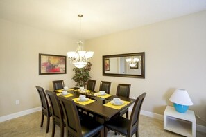 A dining room with a rectangular table set for six, surrounded by six chairs. There is a chandelier above the table, and a mirror on the wall to the right.