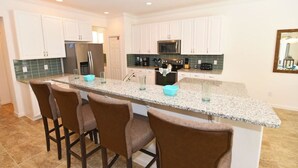 Eine Küche mit weißen Schränken, einer Granit-Arbeitsplatte, einem Edelstahl-Kühlschrank, einer Mikrowelle und einer Kaffeemaschine. Vier braune Barhocker und eine kleine turquoise Schale stehen auf der Arbeitsplatte.