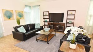 Ein Wohnzimmer mit schwarzem Ledersofa, einem Holztisch, einem TV-Schrank und einem Fernseher. Es gibt zwei Wandregale und einen Topfpflanzen.