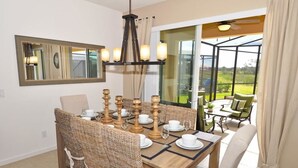 Un coin repas est aménagé avec une table en bois, dressée pour six personnes, et entourée de chaises en osier. Un grand miroir est accroché au mur, au-dessus de la table, et une porte vitrée donne sur un patio extérieur offrant une vue sur la verdure.