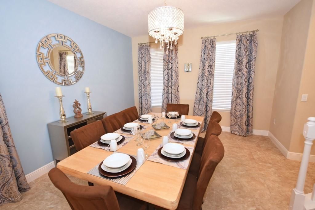 Comedor con una mesa rectangular dispuesta para seis personas, rodeada de sillas marrones. Un candelabro cuelga del techo, y hay un espejo decorativo en la pared.