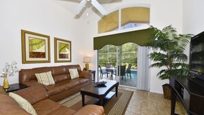 A living room with a brown leather sofa, a coffee table, a TV, and a large window with a view of a pool and greenery. - 4452 Veranda Palms House 4 Bedroom by Florida Star (Kissimmee)