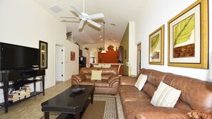 A living room with a brown leather sofa, a black coffee table, a television, and framed botanical prints on the wall. - 4452 Veranda Palms House 4 Bedroom by Florida Star (Kissimmee)