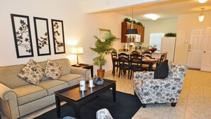 A living room with a sofa, armchair, coffee table, and a dining area with a table and chairs. There is a plant on the coffee table and a lamp on the side table. - 4569 Bella Vida Condo 3 Bedroom by Florida Star (Kissimmee)