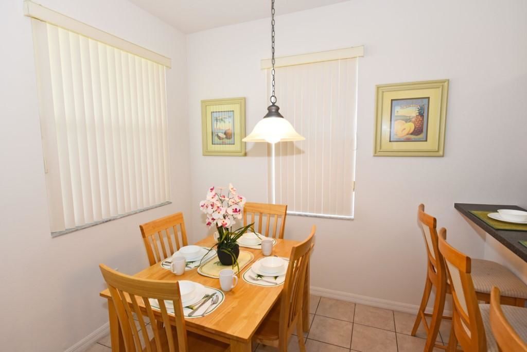 A dining area with a wooden table set for four, surrounded by wooden chairs. There is a hanging light fixture above the table and two framed pictures on the walls.