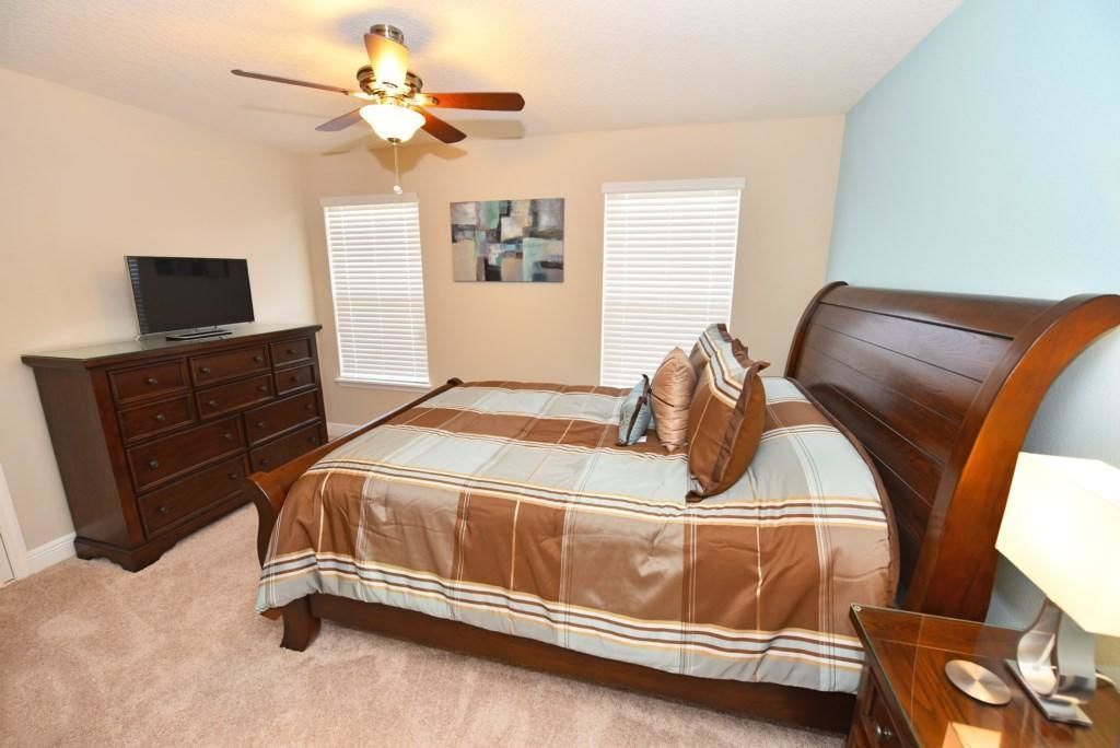 A bedroom with a bed, a dresser, a television, a ceiling fan, and a painting on the wall.