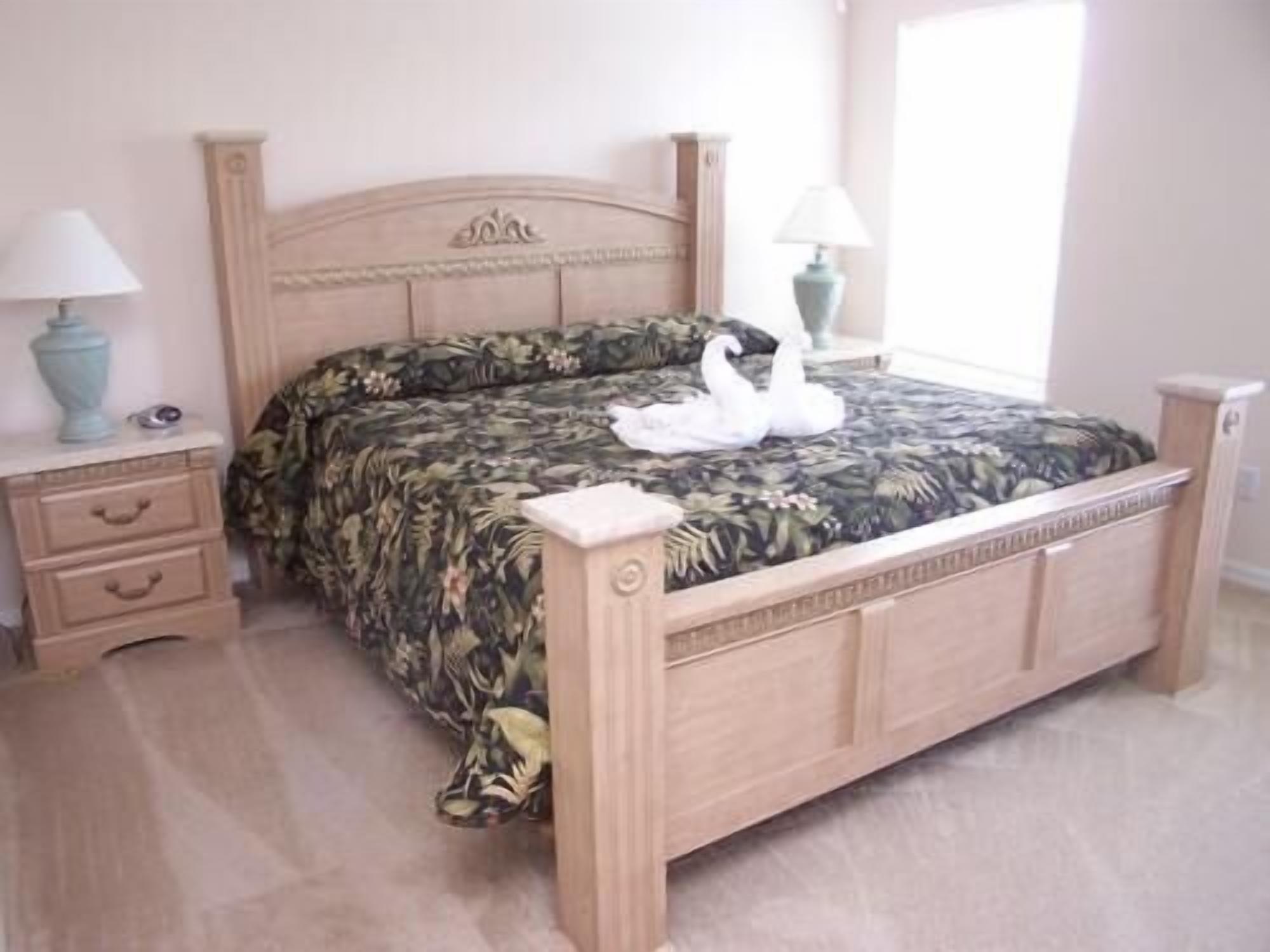 Een slaapkamer met een houten bed, een bloemenprint beddeken, twee nachtkastjes met lampen, en een raam met jaloezieën.