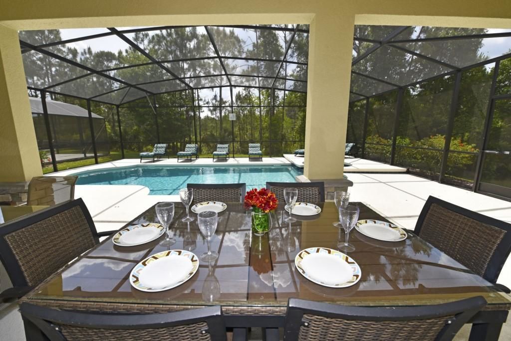 An outdoor dining area with a glass table, wicker chairs, and a pool in the background.