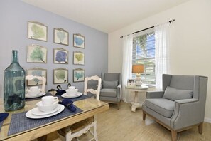 A dining area with a table set for two, surrounded by chairs, a window with blinds, and framed artwork on the wall. - 670 Watersong House 7 Bedroom by Florida Star (Davenport)