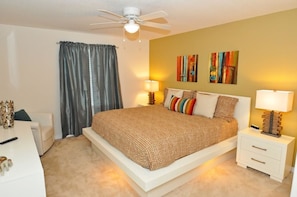 A bedroom with a large bed, two bedside tables with lamps, a ceiling fan, and a window with curtains.