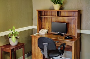 A wooden desk with a computer monitor, a telephone, and a chair. There is a potted plant on a small table beside the desk. - Rodeway Inn (Edson)