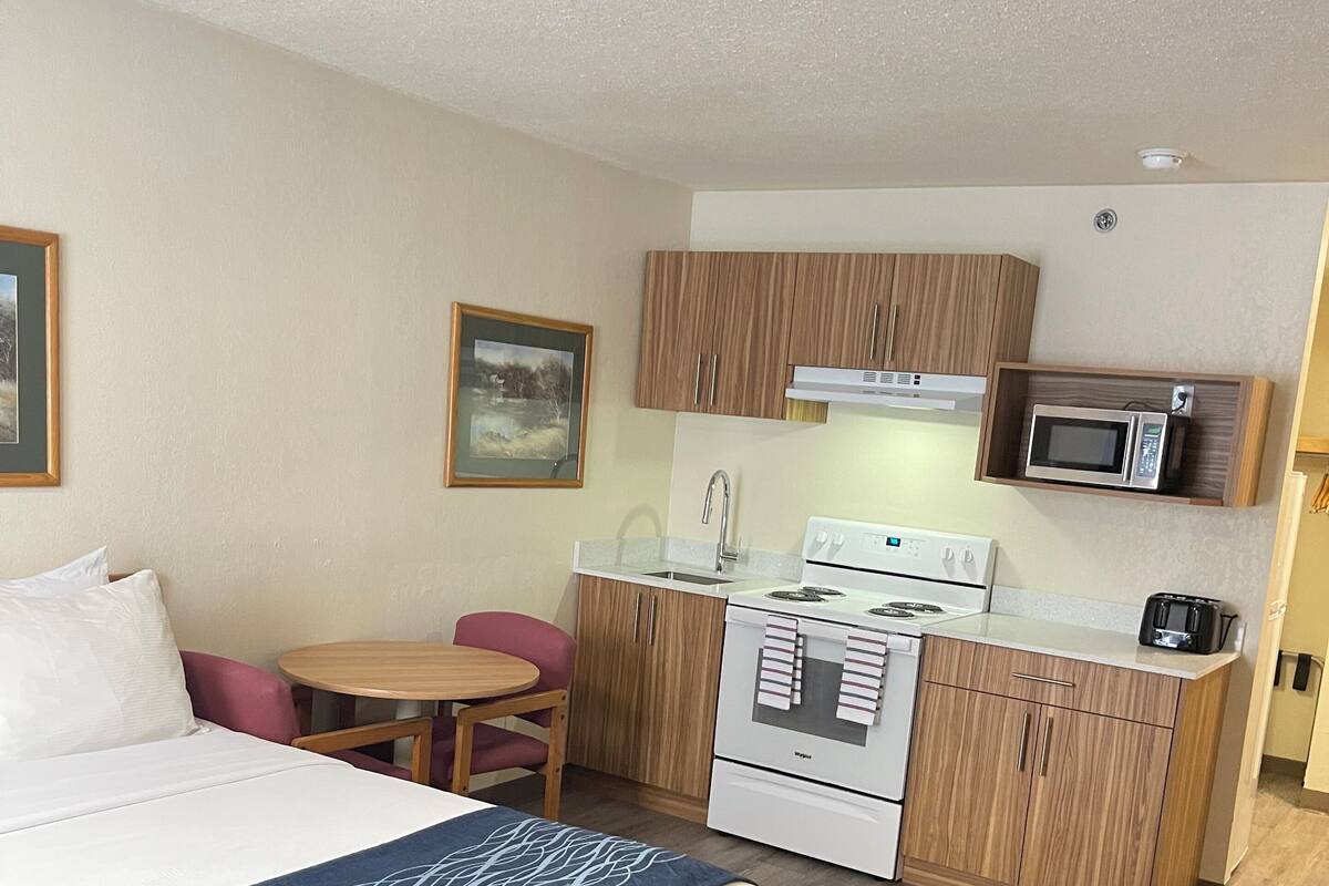 Una habitación de hotel compacta con una cama, una pequeña mesa de comedor y una kitchenette equipada con microondas, estufa y fregadero.