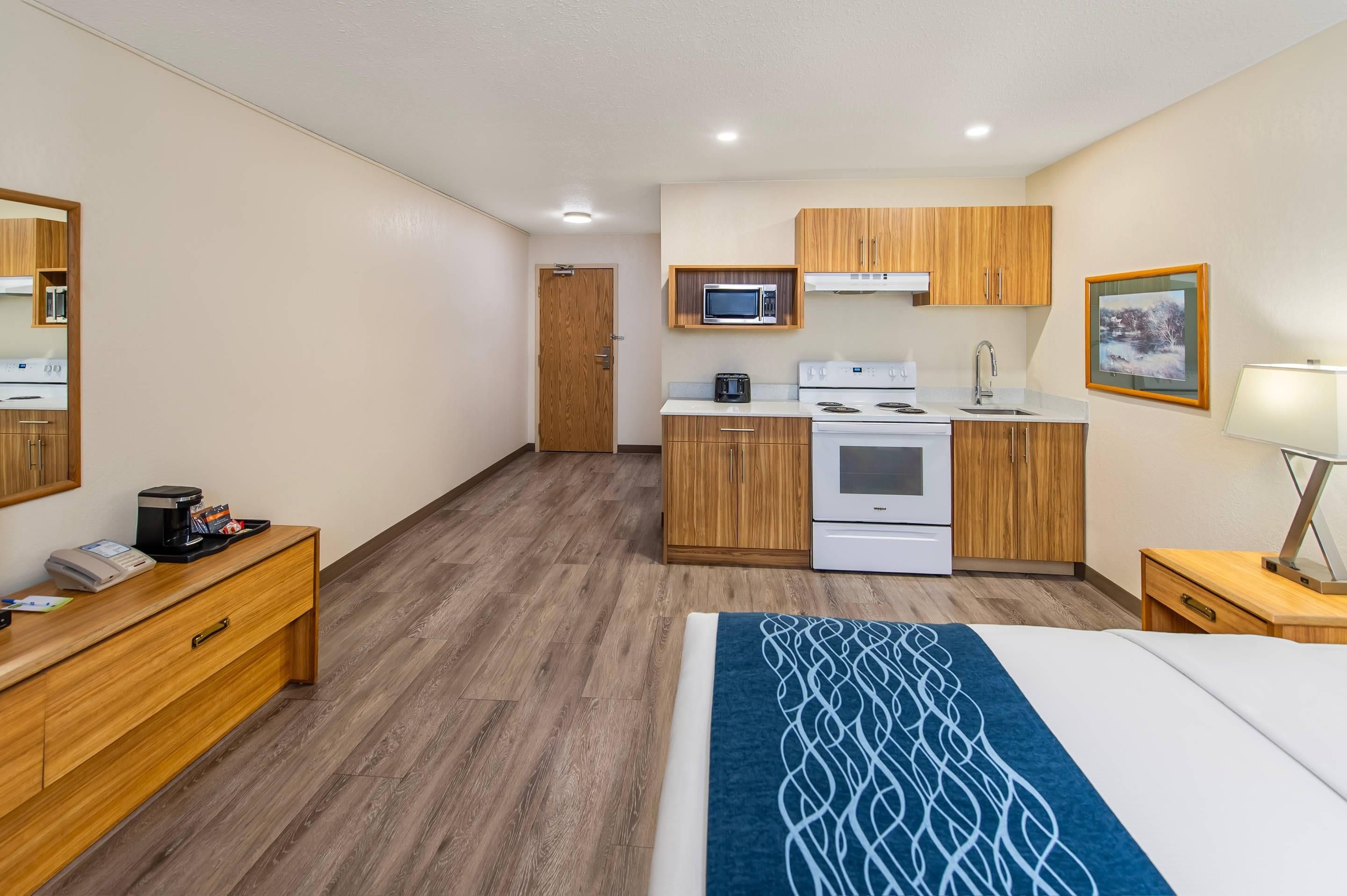Una habitación de hotel compacta con kitchenette, una cama, una mesita de noche con lámpara y un aparador de madera.