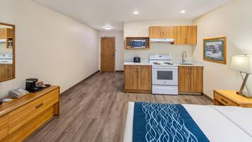 Una habitación de hotel compacta con kitchenette, una cama, una mesita de noche con lámpara y un aparador de madera.