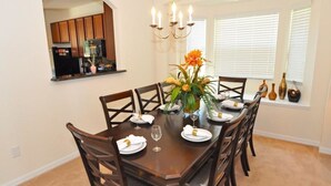 A dining room with a dark wooden table set for four, surrounded by chairs. There is a chandelier above the table, and a window with blinds to the right.