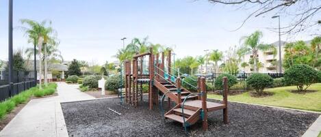 Un parque infantil con tobogán y estructuras para escalar, rodeado de un área ajardinada con árboles y un sendero.