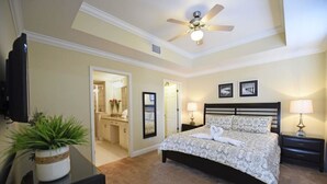 A bedroom with a bed, a ceiling fan, a TV, a desk with a plant, and a bathroom visible through an open door.